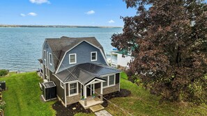 A two-story house with a sloped roof, a front porch, and a view of a lake. - Renovated Lakefront Home- Deck, Grill and Views (Seneca Falls)