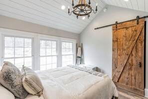 Ein Schlafzimmer mit einem großen Bett, einer Chandelieber, einer Holztür im Scheunenstil und Blick ins Freie durch große Fenster.