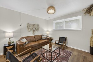En moderne stue med en brun lædersofa, et glasbord, en vævet måtte og et stort vindue med persienner.