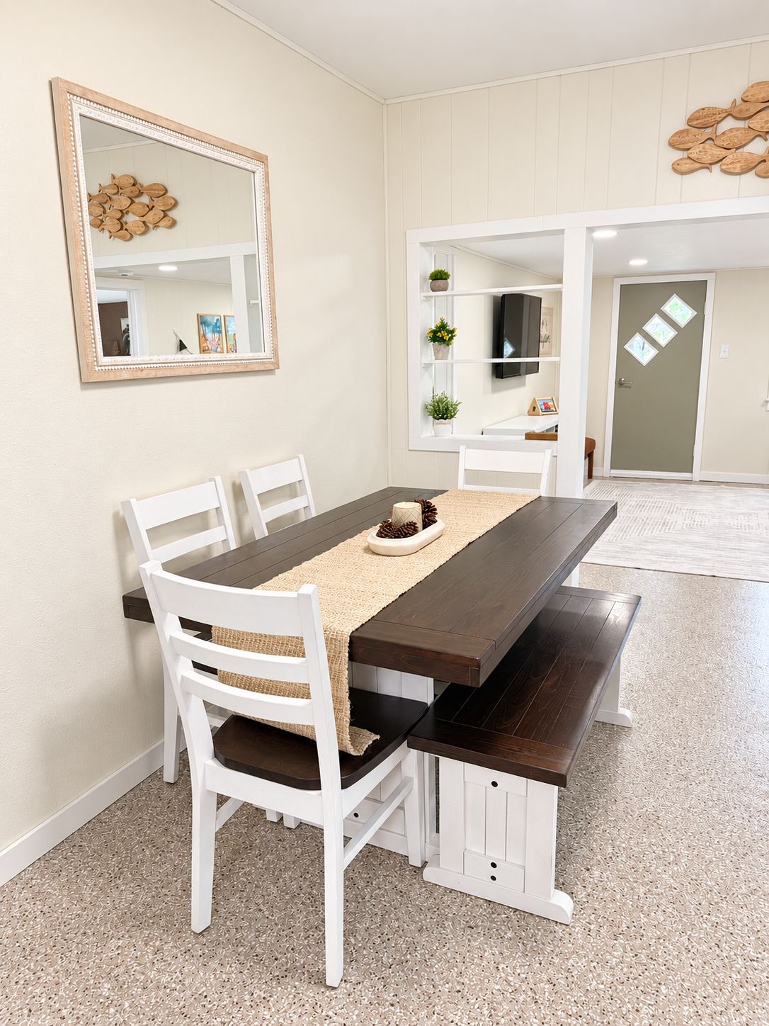 A dining area with a rectangular table, white chairs, and a bench. There is a mirror on the wall, a shelf with plants, and a door with a diamond-shaped window.