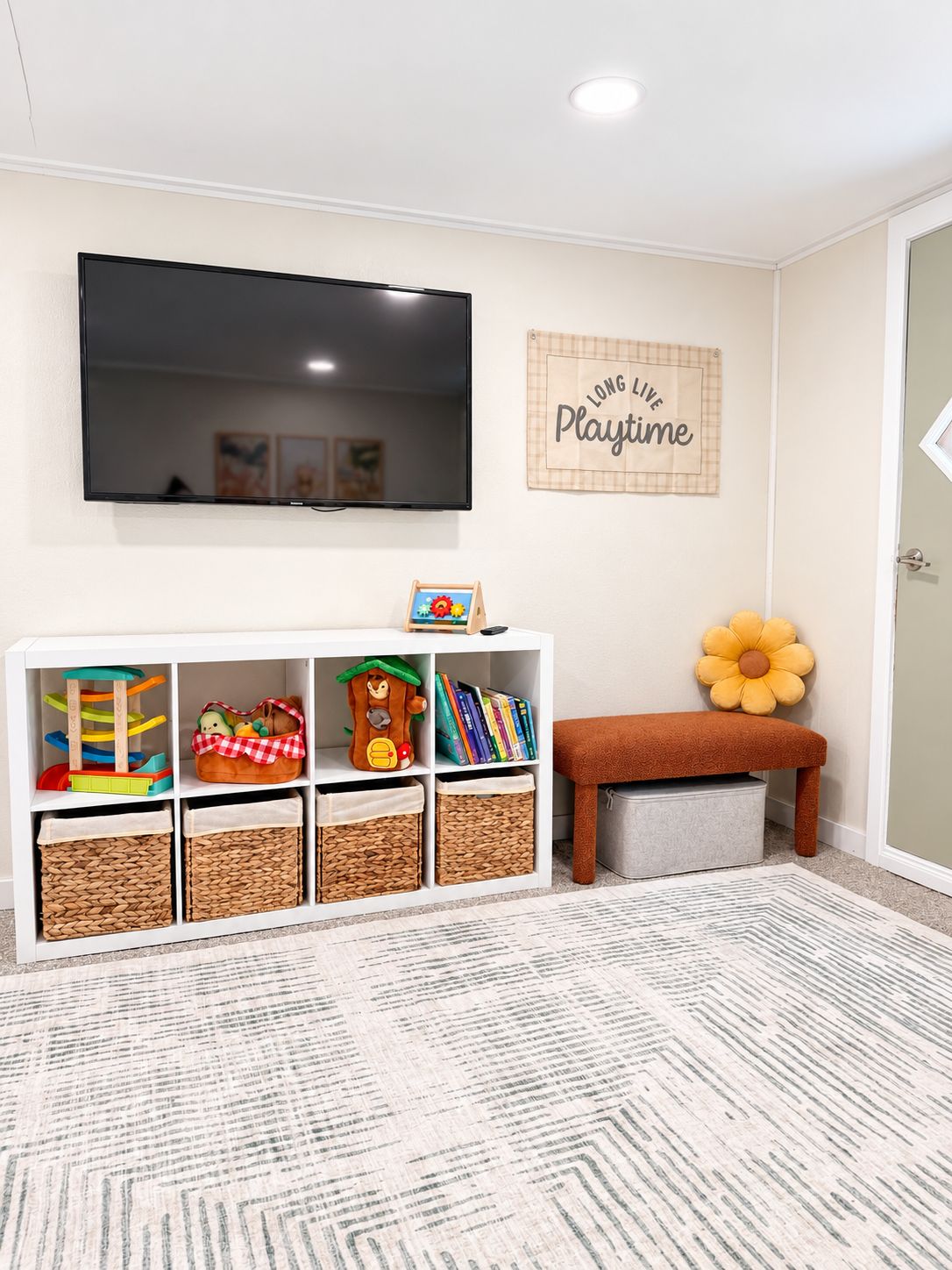 A room with a flat-screen TV mounted on the wall, a bookshelf with toys and books, a bench, and a wall sign that reads 'LONG LIVE Playtime'.