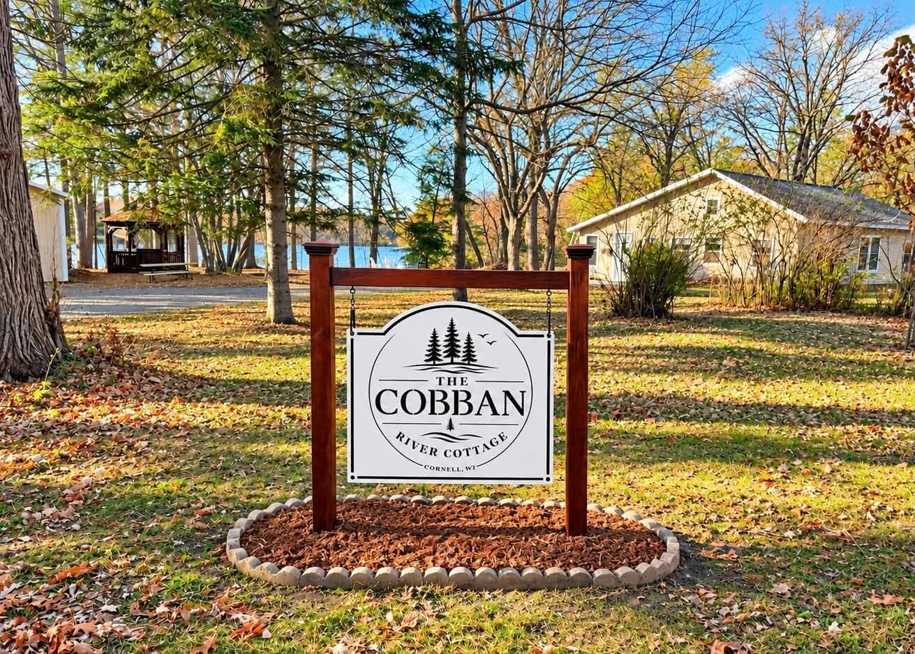 A sign for The Cobban River Cottage in Cornell, WI, surrounded by trees and a grassy area.