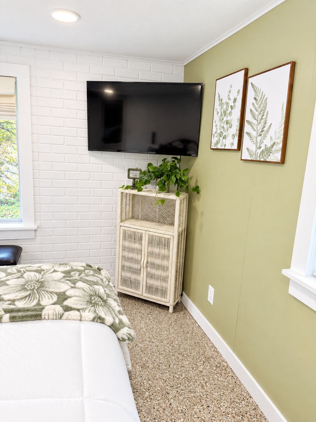 A bedroom with a bed, a television mounted on the wall, a small plant on a side table, and framed botanical prints on the wall.