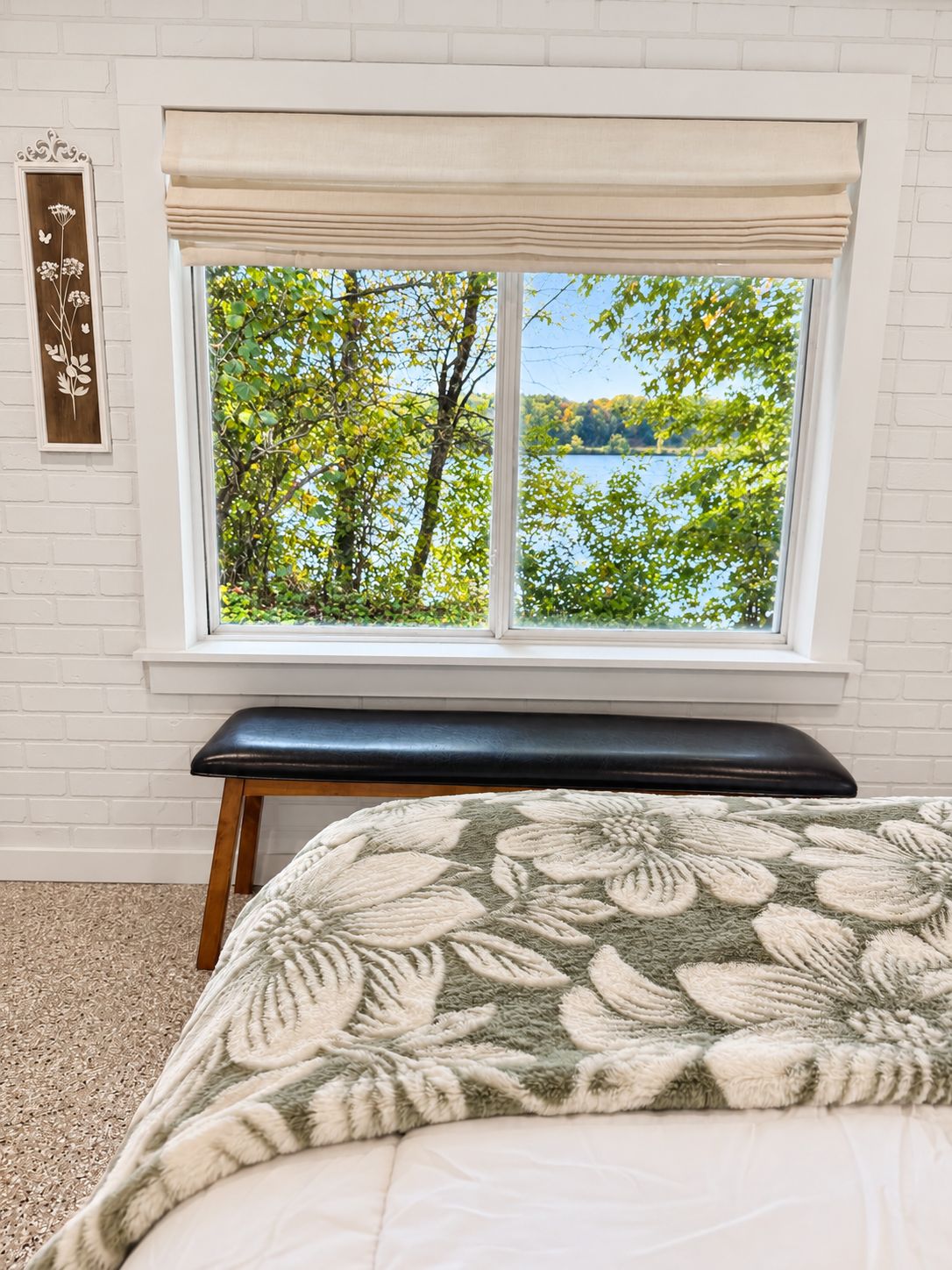 A bedroom with a large window, a bed with a floral-patterned blanket, a bench, and a wall-mounted decoration.