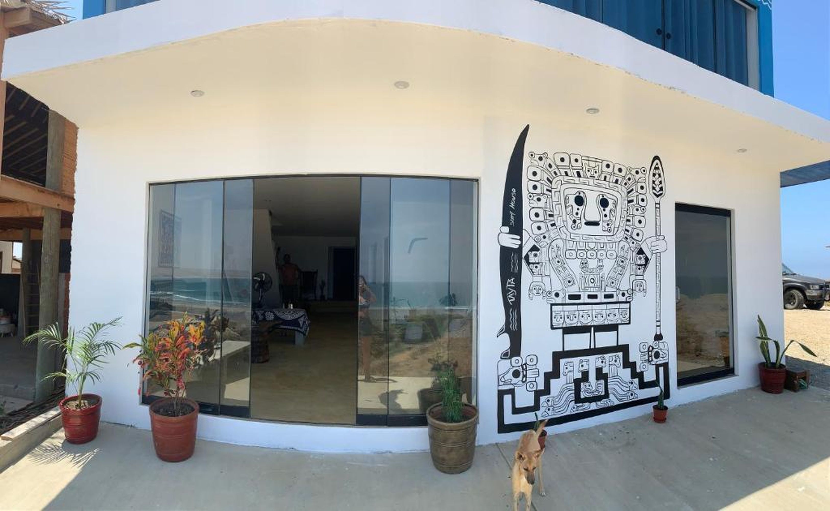 A modern building with large glass doors and windows, a mural on the exterior wall, and potted plants outside.