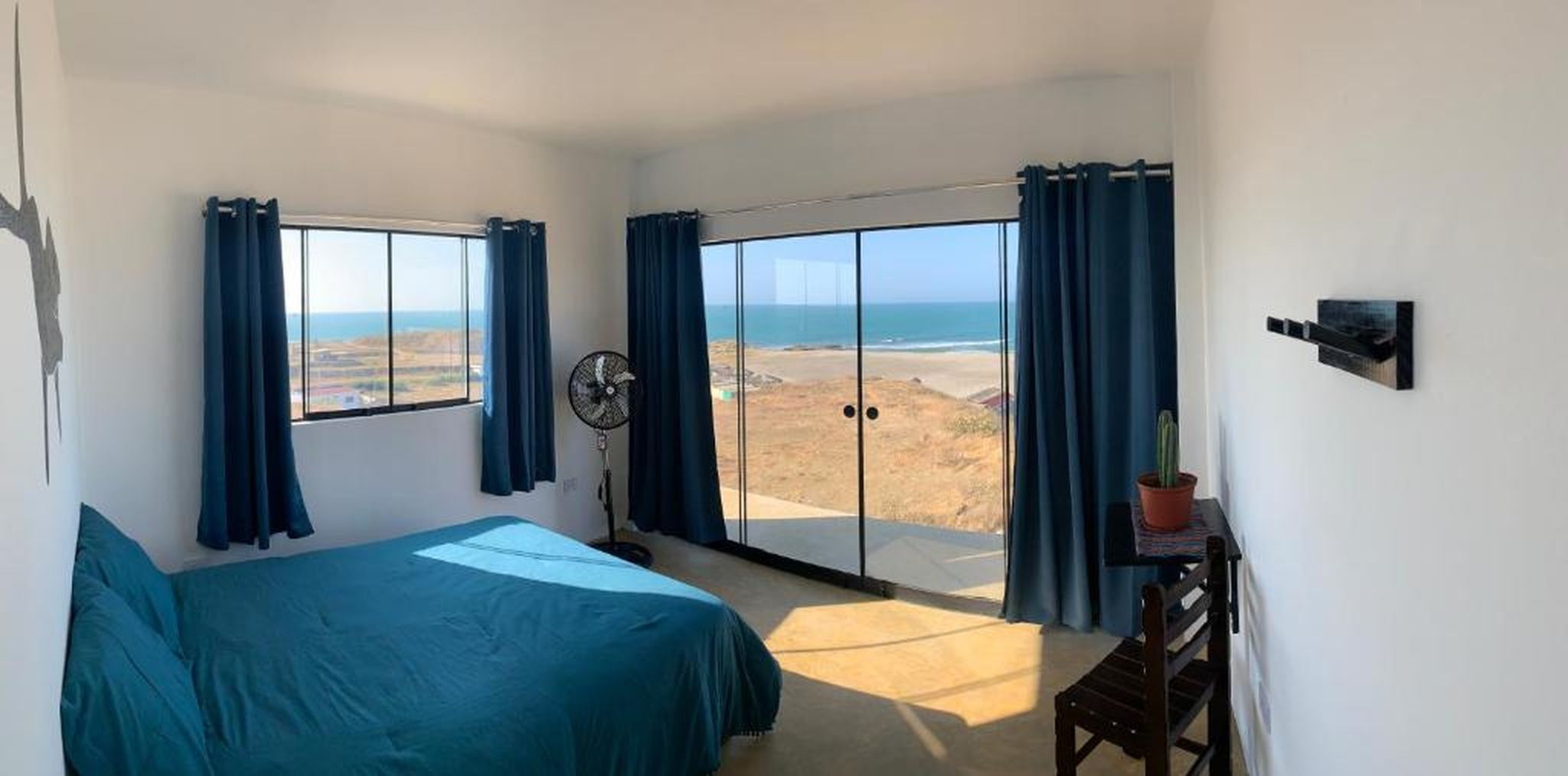A bedroom with a bed, a fan, a potted plant, and a view of the beach through large sliding doors.