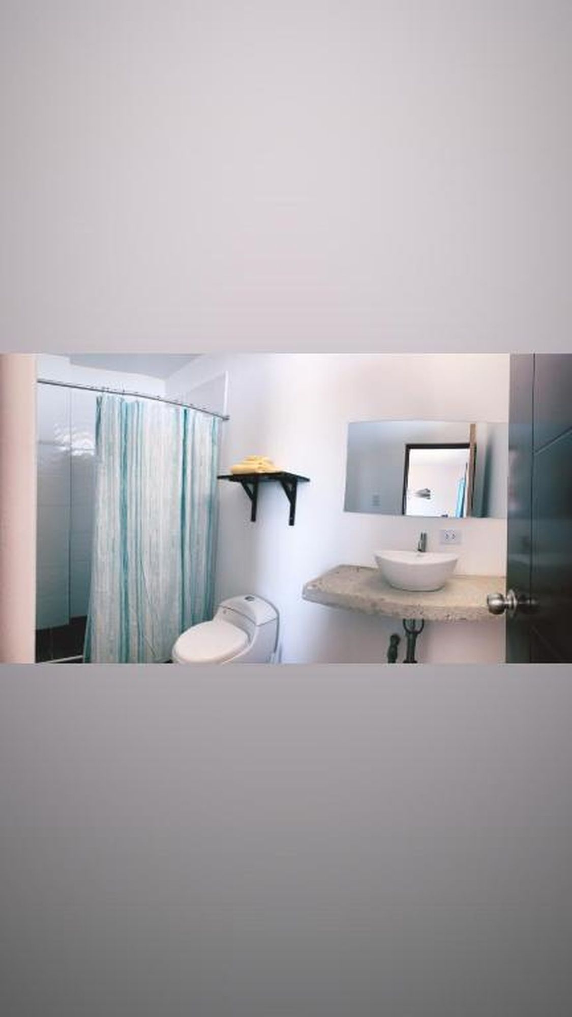 A bathroom with a glass shower enclosure, a toilet, a sink with a mirror above it, and a shelf with a towel.