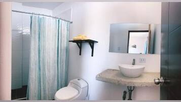 A bathroom with a glass shower enclosure, a toilet, a sink with a mirror above it, and a shelf with a towel.