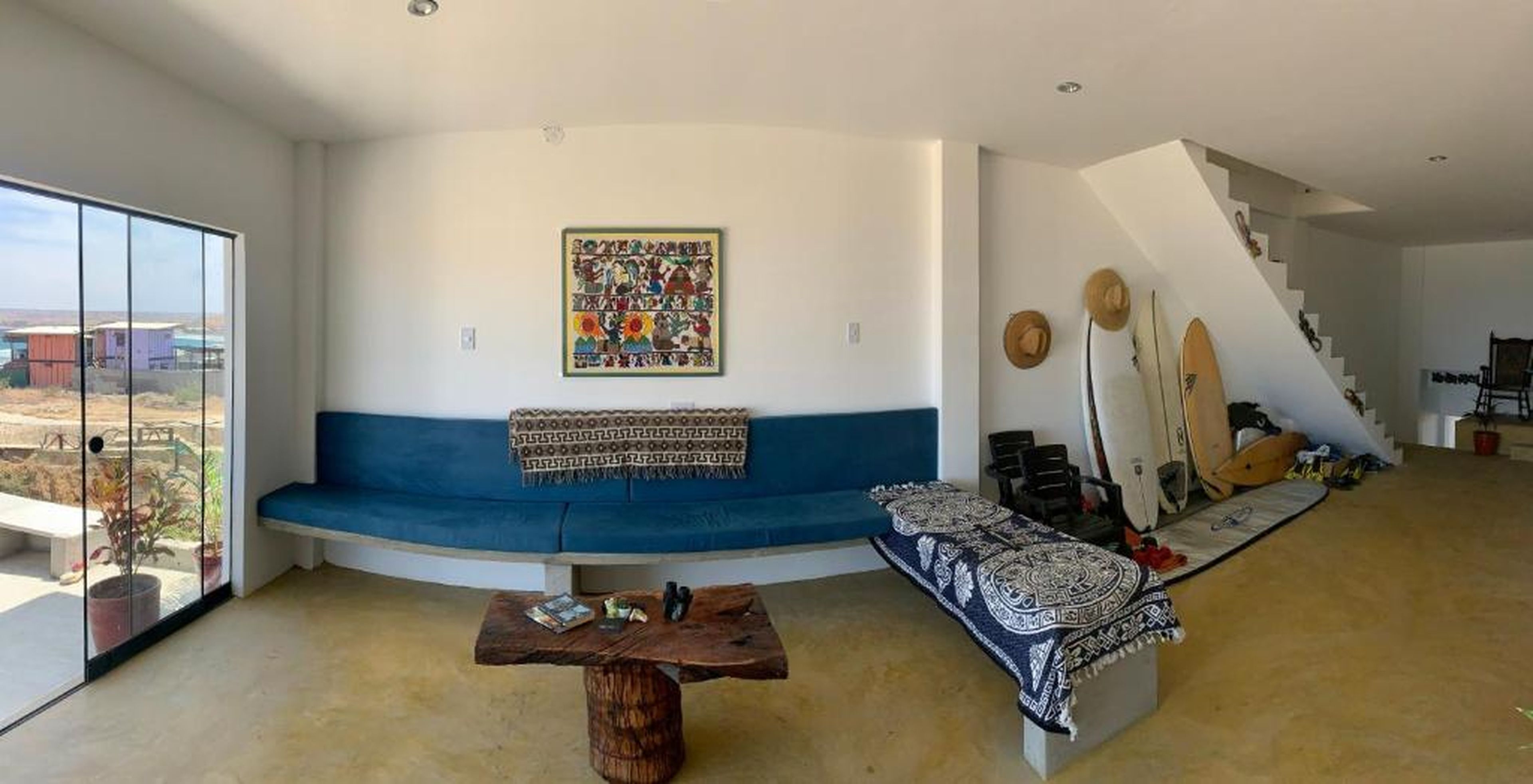 Un salon avec un canapé bleu, une table basse en bois, un tableau abstrait aux couleurs vives et un escalier orné de planches de surf.