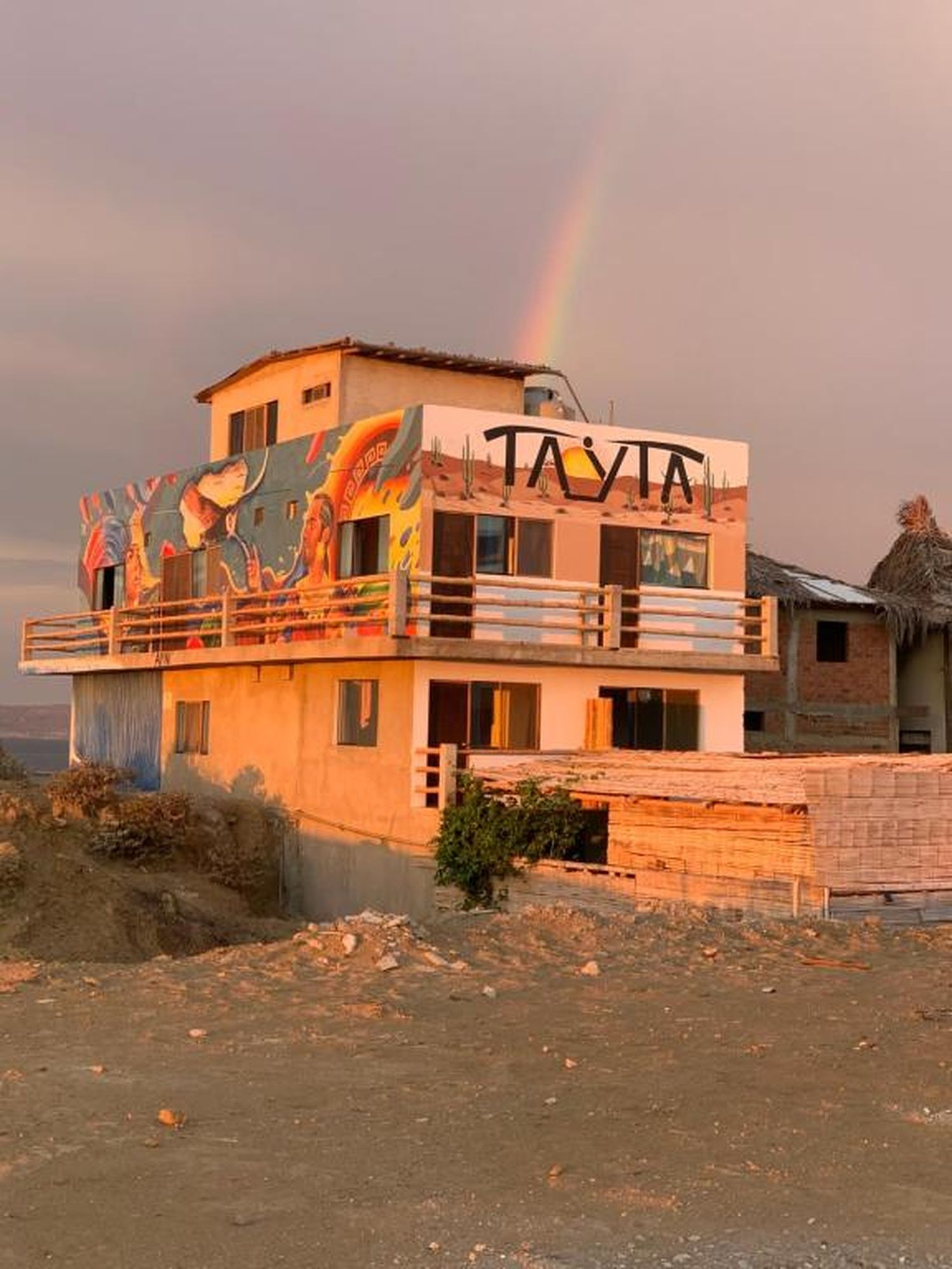 A two-story building with a colorful mural on the side, a balcony, and a thatched-roof structure nearby.
