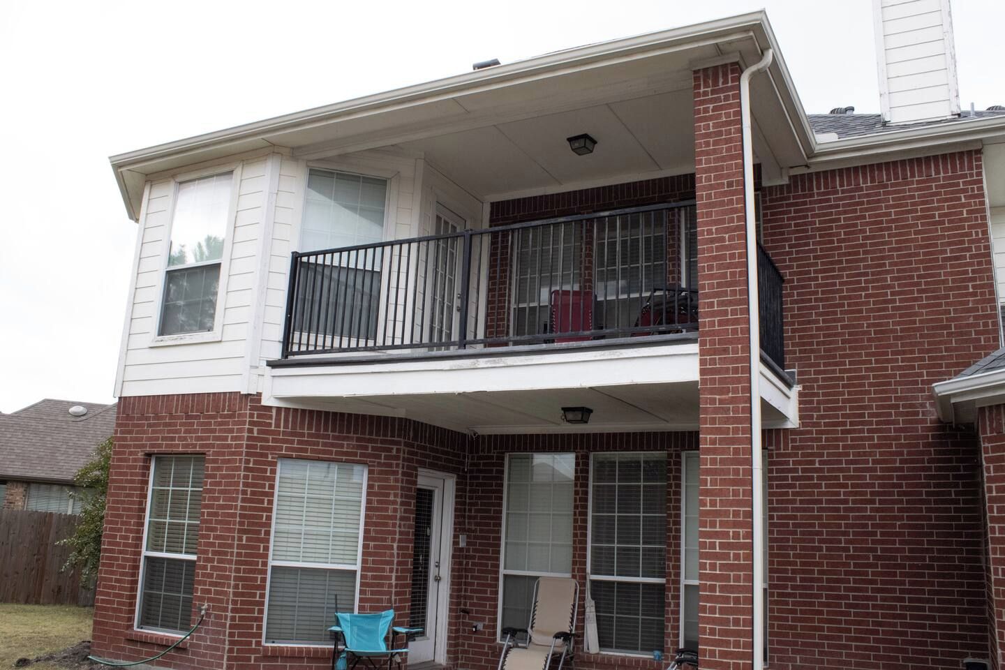 A two-story residential building with a balcony on the upper floor, a front door, and a small porch area with chairs.