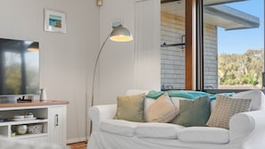 Ein modernes Wohnzimmer mit einem weißen Sofa, einem Flachbildfernseher, einer Stehlampe und Blick durch eine Glastür.