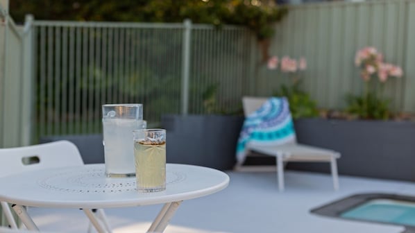 Ein weißer Sitzbereich im Freien mit einem Tisch, zwei Gläsern eines klaren Getränks und einem Metallzaun im Hintergrund.
