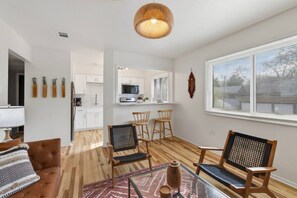 Un salon moderne avec un canapé, des fauteuils, une table basse en verre et une cuisine en arrière-plan.