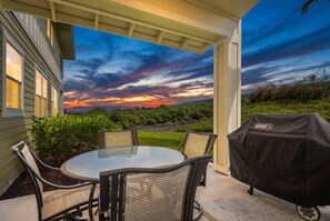 An outdoor patio with a round table and chairs, a grill, and a view of a sunset over a landscape. - Na Hale O Keauhou#n6: Beautiful Corner Townhome! (Kailua-Kona)