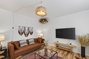 A modern living room with a brown leather sofa, a glass coffee table, a woven pendant light, a wall-mounted flat-screen TV, and a wooden console table. - Cozy Stylish Apt in Omaha`s Medical District! (Omaha)
