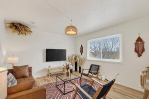 Ein Wohnzimmer mit Flachbildfernseher, einer Couch, zwei Sesseln, einem Couchtisch und einem großen Fenster.