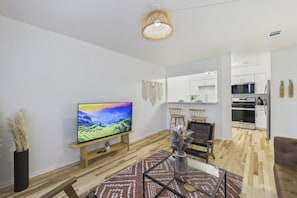Uma sala de estar moderna com televisão de tela plana, piso de madeira, área de jantar com cadeiras e cozinha com armários e eletrodomésticos brancos.