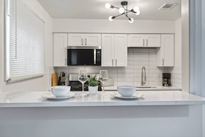 A modern kitchen with white cabinetry, a black microwave, and a central island with two bowls and two wine glasses. - Cozy Stylish Apt in Omaha`s Medical District! (Omaha)