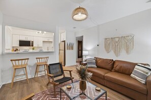 A modern living room with a brown leather sofa, wooden chairs, a glass coffee table, and a kitchen area with white cabinets and a microwave. - Cozy Stylish Apt in Omaha`s Medical District! (Omaha)