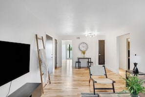 A spacious living room with a wooden floor, a flat-screen TV, a painting on the wall, a dining table with chairs, and a clock. - Prime Location! HGTV Design! (Omaha)