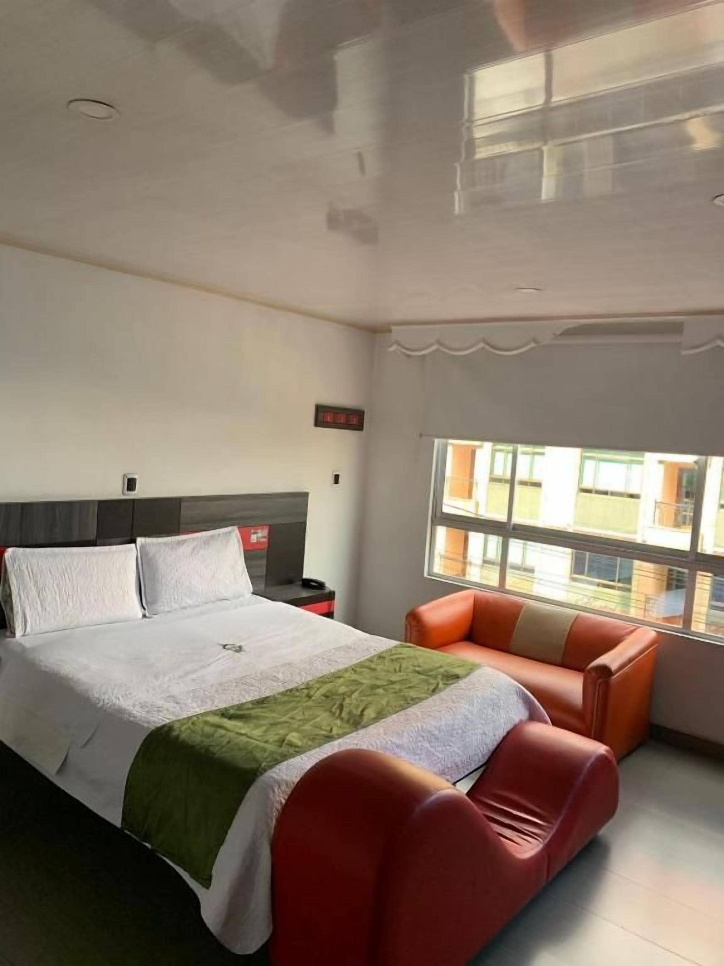 A hotel room with a bed, a red sofa, a window with a view, and a digital clock.