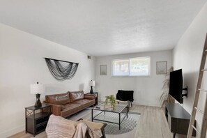 A modern living room with a brown leather sofa, a coffee table, a TV, and a window with blinds. - Prime Location! HGTV Design! (Omaha)