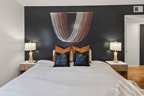 A modern bedroom with a large bed, two bedside tables with lamps, and a unique wall hanging above the bed. - Designer Mid-Term Living Near Omaha Hospitals (Omaha)