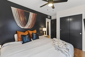 A bedroom with a bed, pillows, a hanging textile, a ceiling fan, and a door. - Designer Mid-Term Living Near Omaha Hospitals (Omaha)