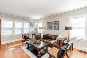 A spacious living room with a brown leather sofa, a wooden coffee table, and a large window with blinds. - Spacious Dundee Duplex | 8BR | Walk to Cafés (Omaha)