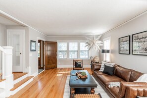 A spacious living room with hardwood flooring, a brown leather sofa, a coffee table, and a large window with natural light. - Spacious Dundee Duplex | 8BR | Walk to Cafés (Omaha)