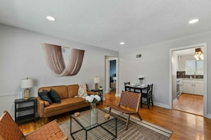A living room with a sofa, coffee table, and chairs, a dining area with a table and chairs, and a kitchen with cabinets and a window. - Prime Location Midtown Omaha! (Omaha)