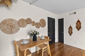 A dining area with a wooden table, chairs, and a vase of flowers. There are decorative wall hangings and a door in the background. - Designer Mid-Term Living Near Omaha Hospitals (Omaha)