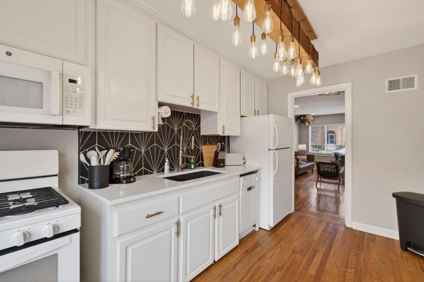 A modern kitchen with white cabinetry, a built-in microwave, a stove, and a refrigerator. There is a wooden floor and a hanging light fixture.