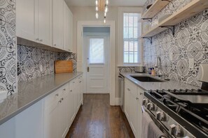 A kitchen with white cabinets, patterned backsplash, a wooden cutting board, a stove, and a window with blinds. - Storybook Retreat | Walk to Dining & Coffee (Omaha)