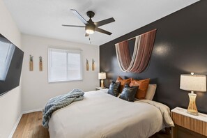 A bedroom with a bed, a ceiling fan, a window with blinds, a wall-mounted lamp, and a decorative wall piece. - Designer Mid-Term Living Near Omaha Hospitals (Omaha)
