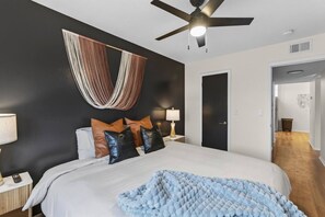 A bedroom with a bed, bedside tables, a ceiling fan, and a hanging textile decoration. - Designer Mid-Term Living Near Omaha Hospitals (Omaha)