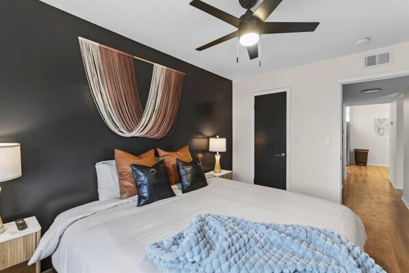 A bedroom with a bed, bedside tables, a ceiling fan, and a hanging textile decoration. - Designer Mid-Term Living Near Omaha Hospitals (Omaha)