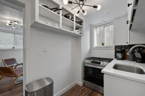 Une cuisine moderne avec des meubles blancs, une cuisinière en acier inoxydable et un four encastré. Une fenêtre avec des stores et une petite plante sur le rebord de la fenêtre complètent l’ensemble.