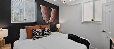 A bedroom with a large bed, a ceiling light fixture, a window with blinds, and a door.