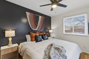 A bedroom with a bed, two bedside tables, a ceiling fan, a window with blinds, and a wall-mounted light fixture. - Designer Mid-Term Living Near Omaha Hospitals (Omaha)