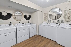 Una sala de bugaderia amb quatre rentamàquines de càrrega frontal blanques, dos miralls fixats a la paret i elements decoratius penjats a la paret.