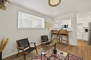 A modern living room with a glass coffee table, two wooden chairs, a dining area with a small table and chairs, and a kitchen with white cabinets and stainless steel appliances. - Cozy Stylish Apt in Omaha`s Medical District! (Omaha)
