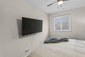 A bedroom with a flat-screen TV mounted on the wall, a window with blinds, and a bed with a grey blanket. - Designer Mid-Term Living Near Omaha Hospitals (Omaha)