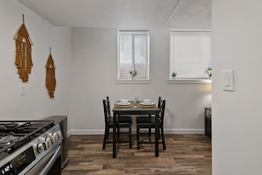 Cozinha compacta com fogão a gás, mesa de jantar com quatro cadeiras e duas janelas com persianas.