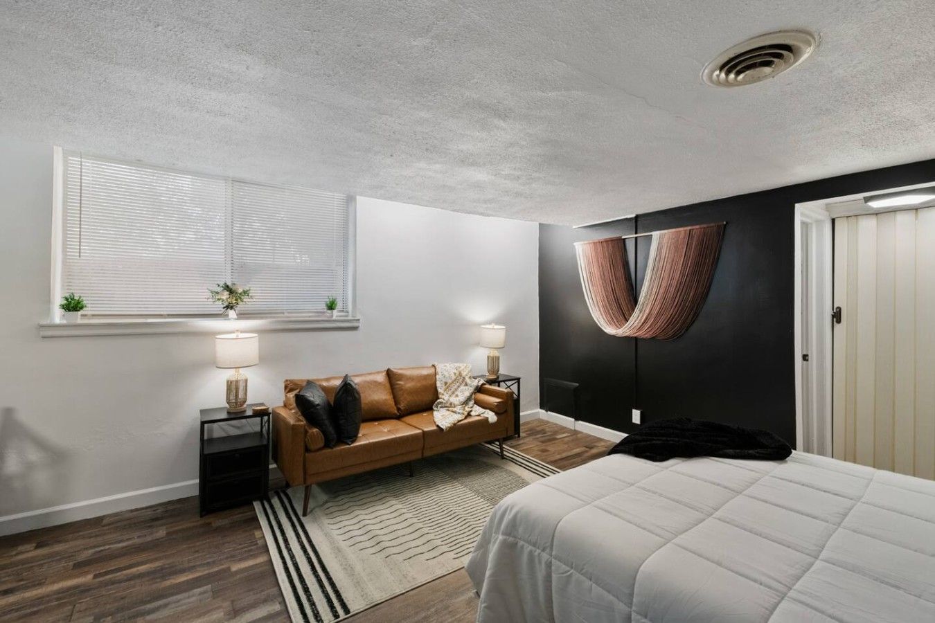 A modern bedroom with a bed, a sofa, a window with blinds, a hanging chair, and a door.