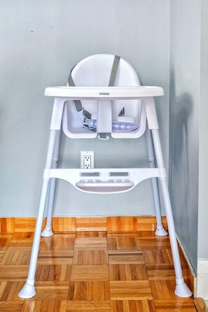 A white child’s high chair with a tray table and a footrest, placed against a wall with a wooden floor. - Comfy Getaway - Walk to West Ed Mall (Edmonton)