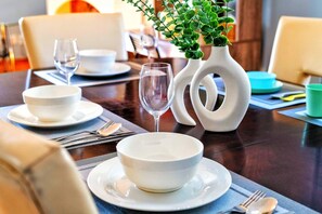A dining table set with white bowls, plates, and cutlery, accompanied by a glass of water and a vase with green plants. - Comfy Getaway - Walk to West Ed Mall (Edmonton)