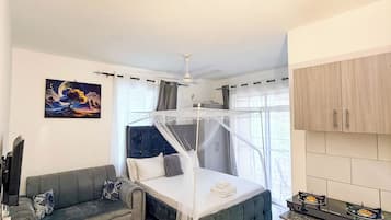 Habitación de hotel compacta con cama, sofá, mesita, televisor y kitchenette.