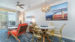 Habitación de hotel con una mesa de comedor redonda, sillas, un ventilador de techo, un ventanal grande y un cuadro de un coche antiguo.